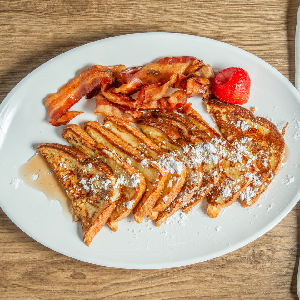 French toast with powdered sugar, bacon, and a strawberry on a white plate with a fork.