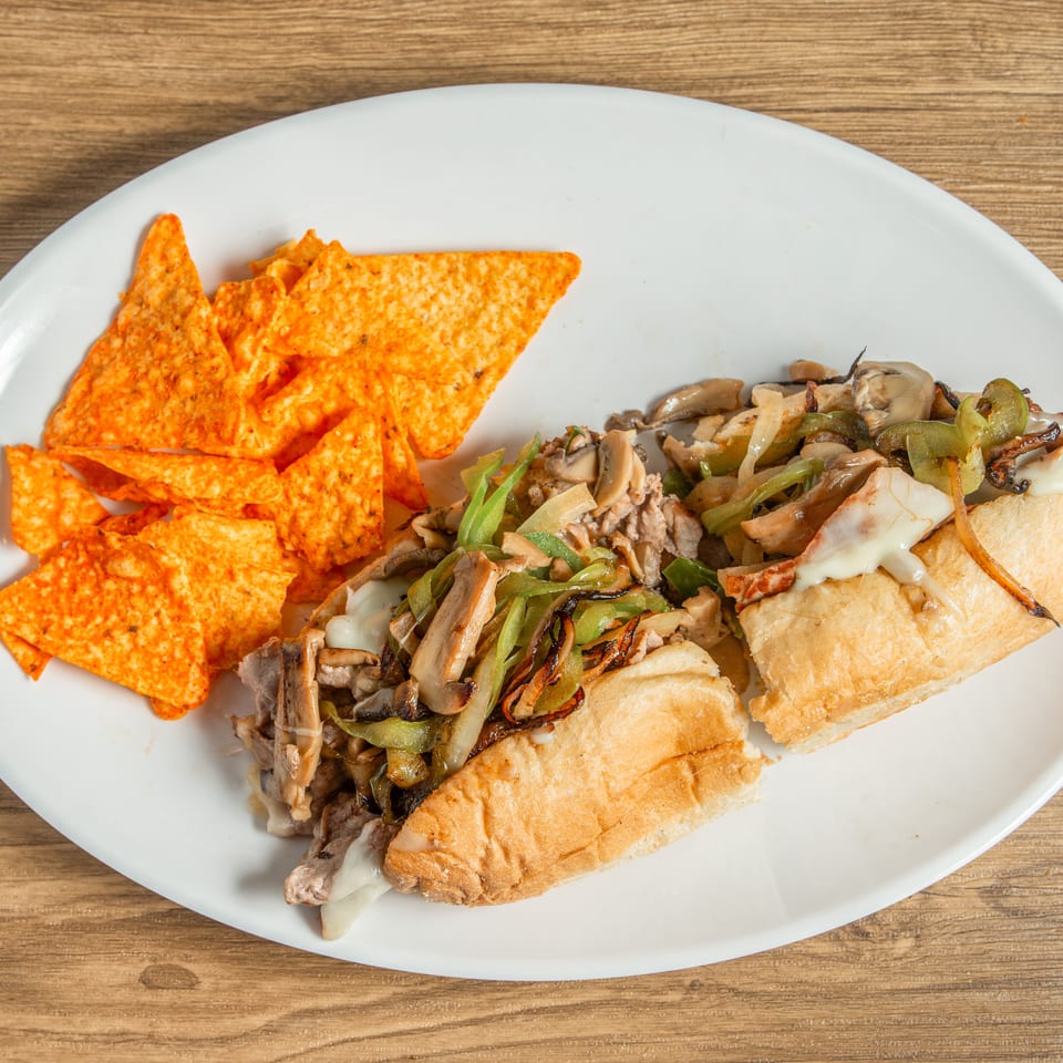 Cheesesteak sandwich with melted cheese, mushrooms, onions, and peppers, served with tortilla chips.
