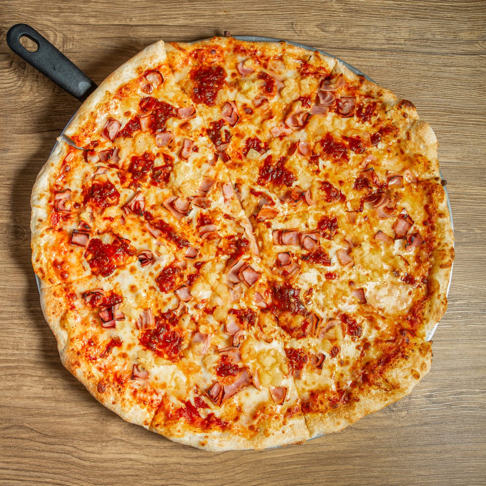 A large, freshly baked Hawaiian pizza with ham and pineapple on a wooden table.